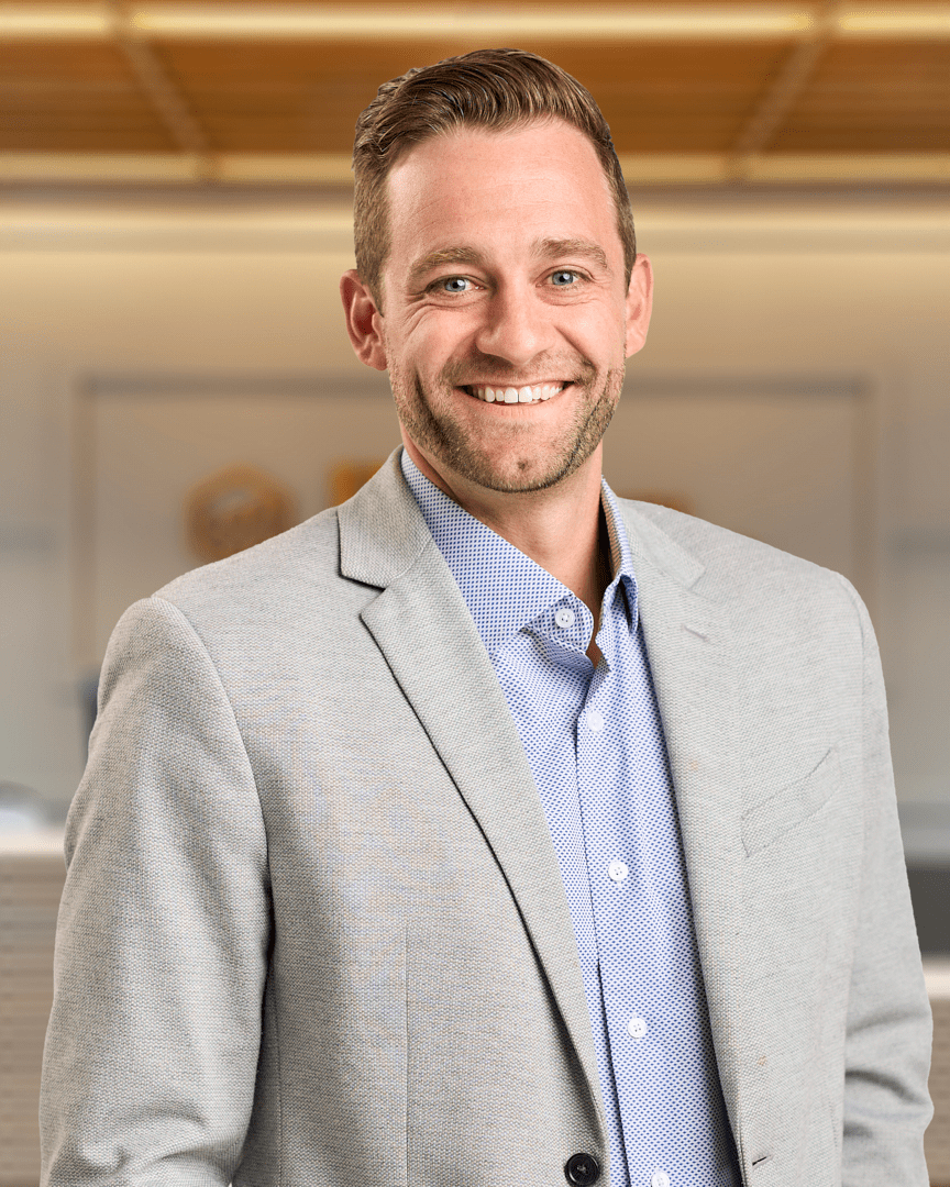 Nate, a man with short brown hair and light stubble smiling in a light grey blazer over a blue patterned shirt, professional headshot with a warm lit office background.