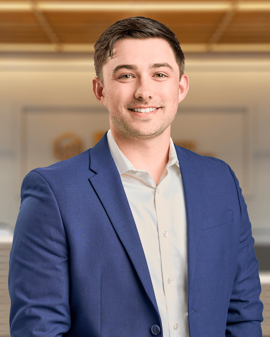 Shane, a man with short brown hair smiling in a navy blazer over a light button up shirt, professional headshot with a warm lit office background.