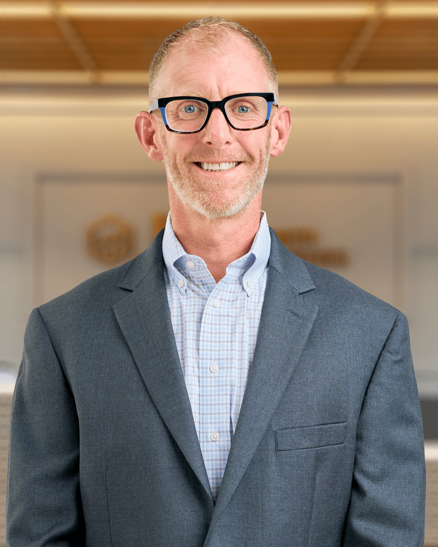 Professional headshot of a man with short gray hair, glasses, and a friendly smile, wearing a gray suit jacket, light blue checkered dress shirt, and blue textured tie, standing indoors against a blurred office background with wood ceiling accents and a gold circular logo visible behind him.