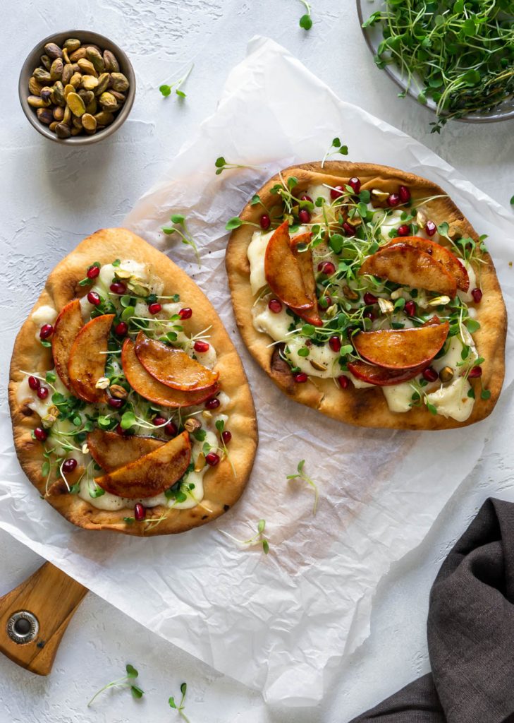 two flatbread pizzas with, pistachios in a small bowl, and micro greens on a plate