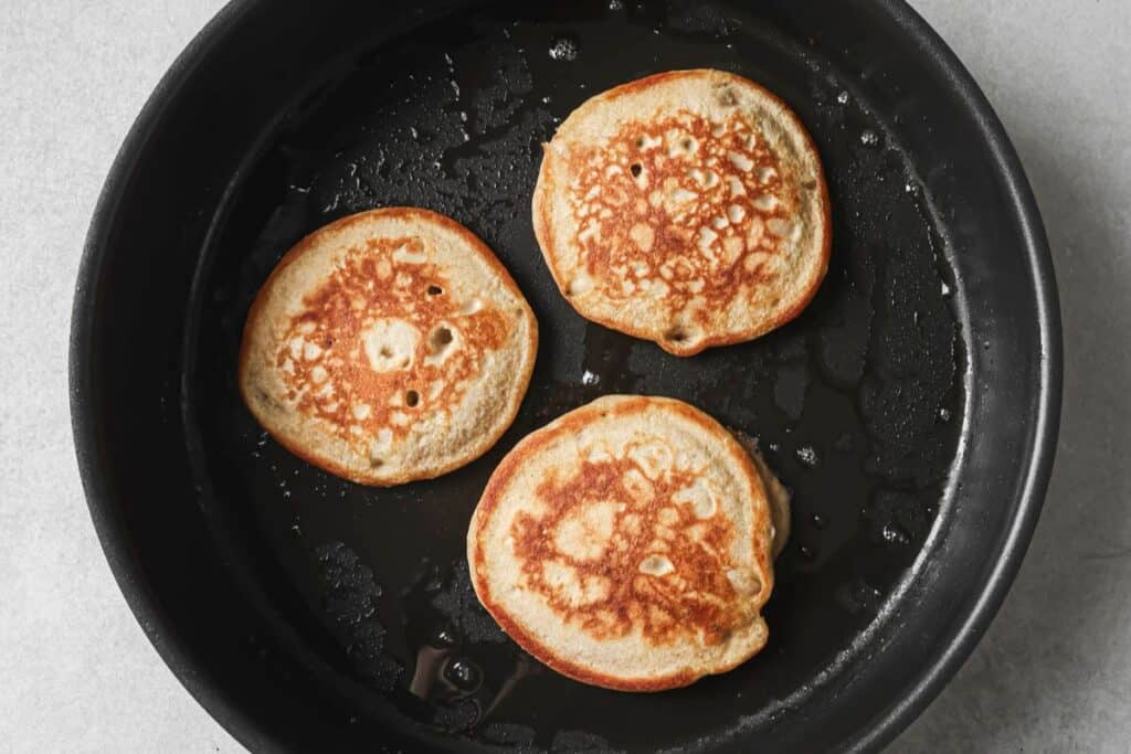 Three pancakes in a skillet browned on the top side. 