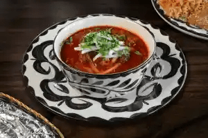 A bowl of red soup topped with chopped onions and cilantro sits on a decorative black-and-white plate. The soup appears rich and garnished, and part of a dish with rice and another with foil are partially visible beside it on a dark wooden table.
