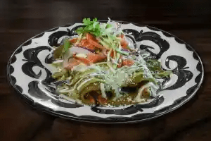 A patterned plate holds a traditional Mexican dish with strips of green nopales (cactus), topped with crumbled cheese, chopped tomato, onion, cilantro, and shredded lettuce on a dark wooden surface.