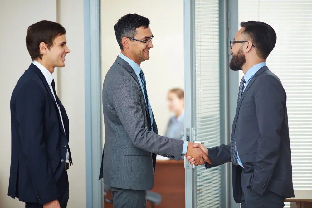 Two businessmen shaking hands in an office to finalize a deal, while a colleague smiles and looks on.