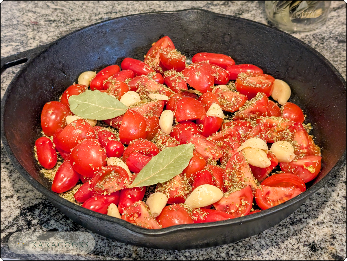 A cast iron skillet on a granite countertop filled with quartered Roma and cherry tomatoes, whole peeled garlic cloves, two bay leaves, and a generous sprinkle of dried oregano, ready to go into the oven.