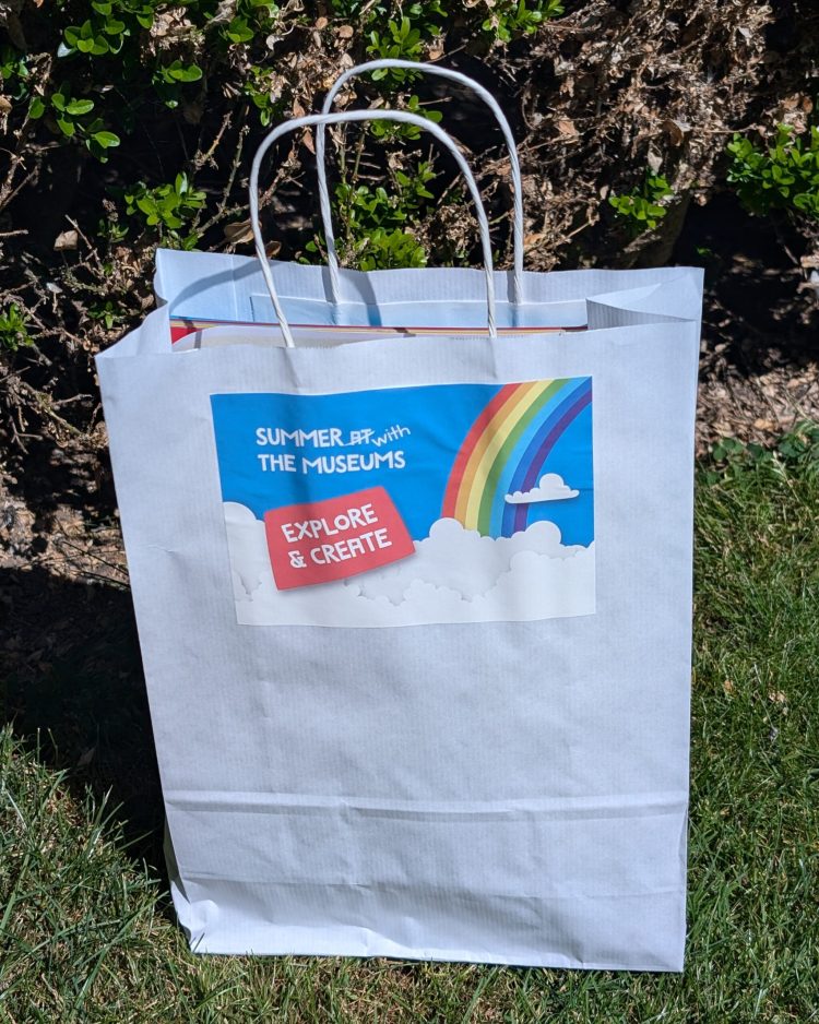A white goody bag with the text "Summer with the Museums, explore & create" featuring a graphic of a rainbow and clouds.