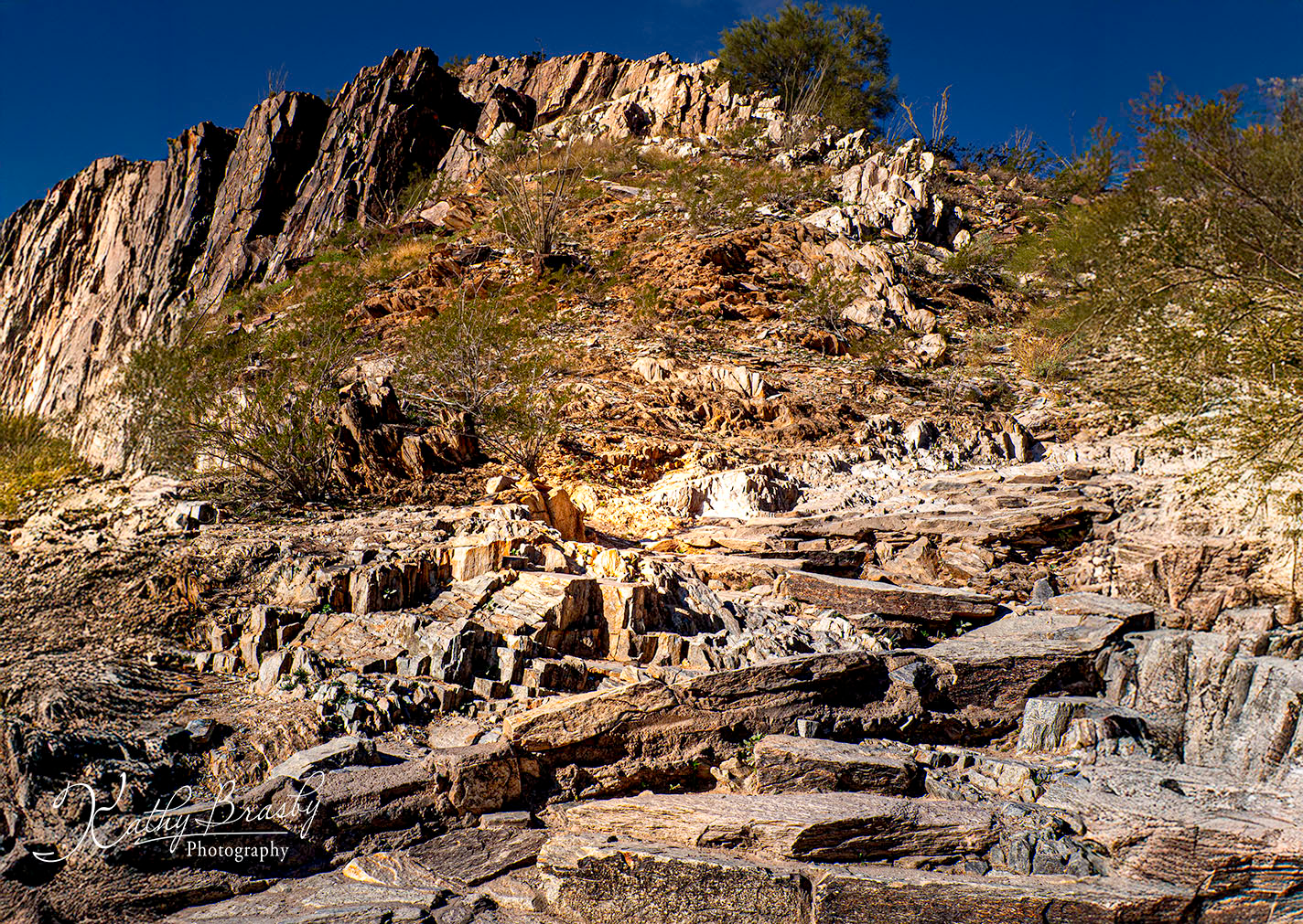 A view looking up a steep, rocky mountainside under a deep blue sky. A challenging path of jagged, uneven stone steps winds up the sunlit slope, dotted with sparse desert bushes.