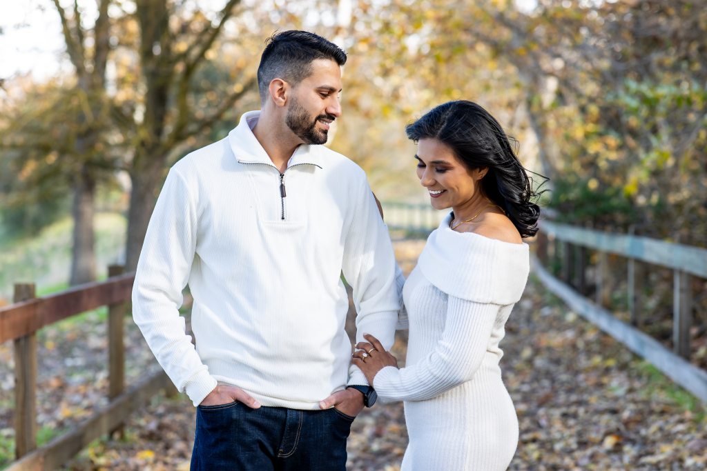 Couple embracing in coordinated white sweaters on a leaf-covered path beside a wooden fence during an autumn engagement shoot. Central Valley photography.