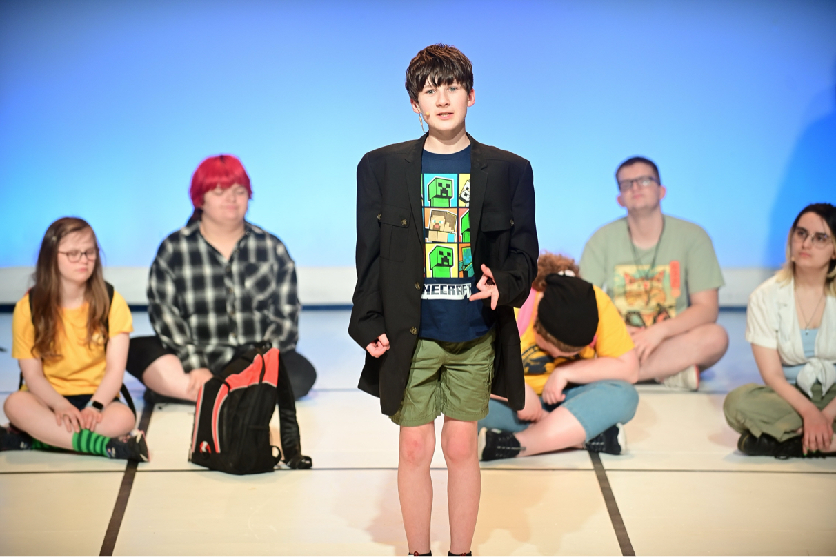 A young teen boy in a blazer speaks in front of other teens.