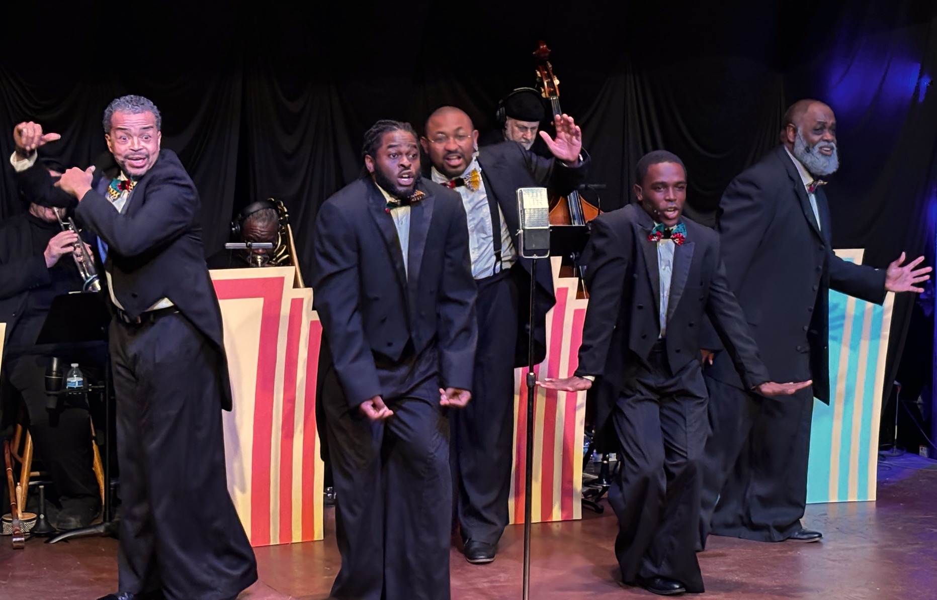 Five Black men in tuxedos dancing in front of a small band.