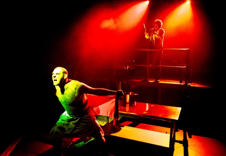 A man playing Bat Boy lunges porward, cast in dramatic red lighting, while another man stands behind him on a platform, with an evil grin.