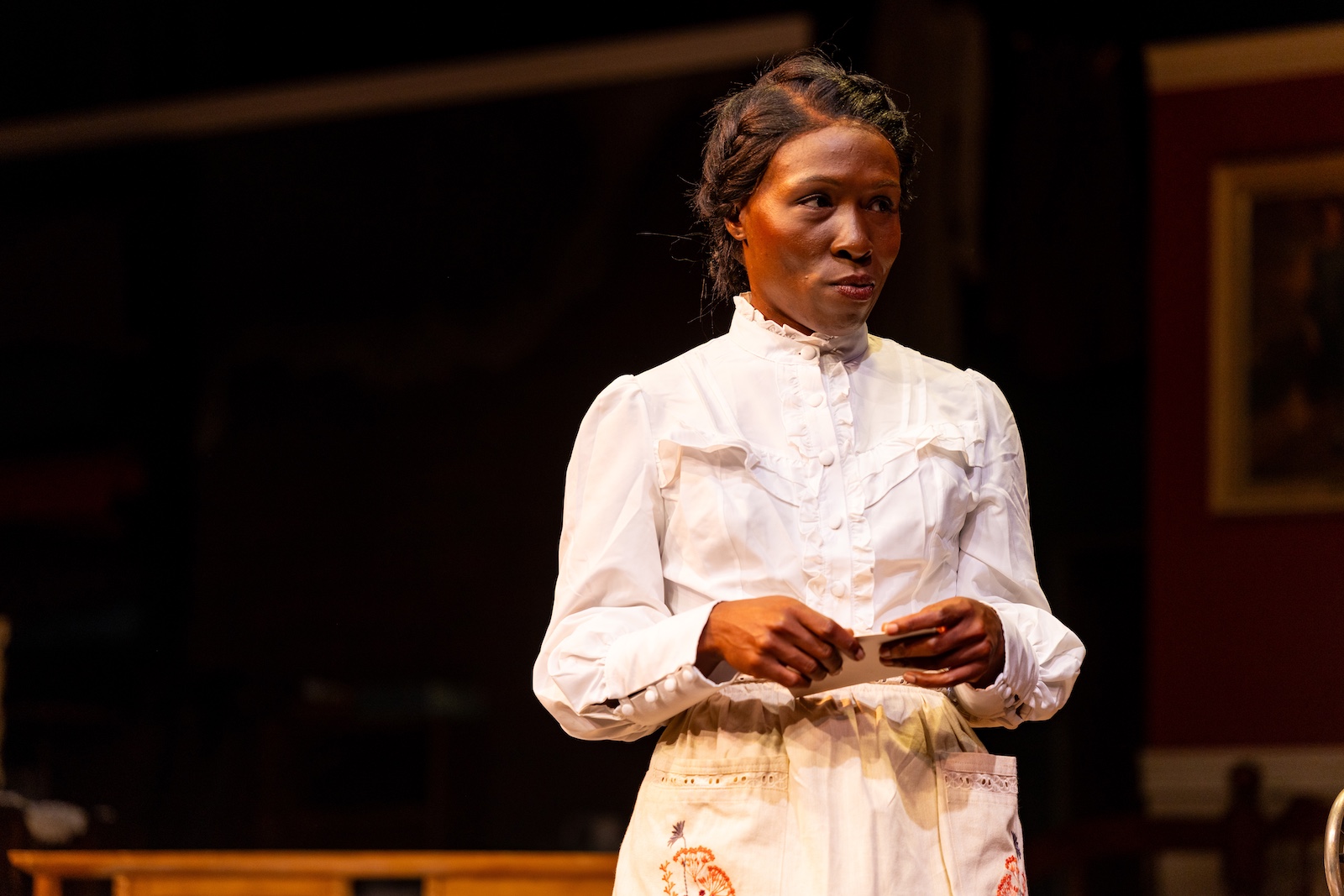 A Black woman in a period costume (1905) stands holding. a letter with a small smirk.