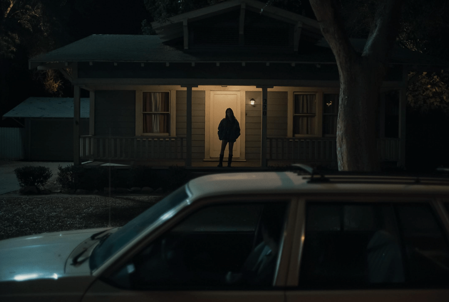 A lone young woman stands on the porch of a suburban house at night while a car is parked in the foreground, creating an eerie, suspenseful scene.