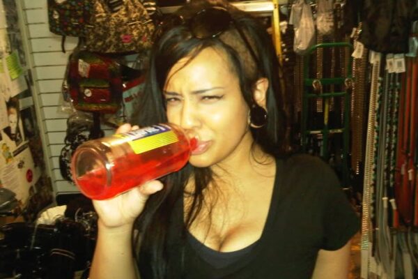 andid photo of a woman with long black hair and winged eyeliner drinking from a red bottle inside a Hot Topic store, surrounded by belts, bags, and accessories.