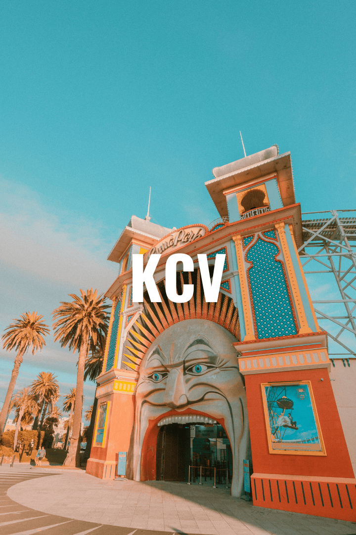 Entrance of an amusement park with a colorful face sculpture and the letters 'KCV' in bold white font overlaying the image, palm trees in the background, under a clear blue sky.