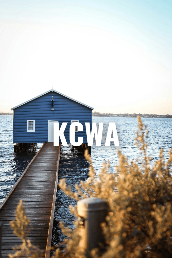 A blue boathouse on stilts over water with a wooden dock leading to it, in a serene waterside setting at sunset or sunrise, with the text 'KCWA' overlaid on the image.
