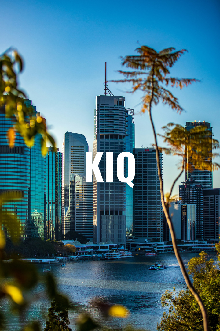 Skyscrapers of a modern city skyline with water in the foreground and trees partially obscuring the view.