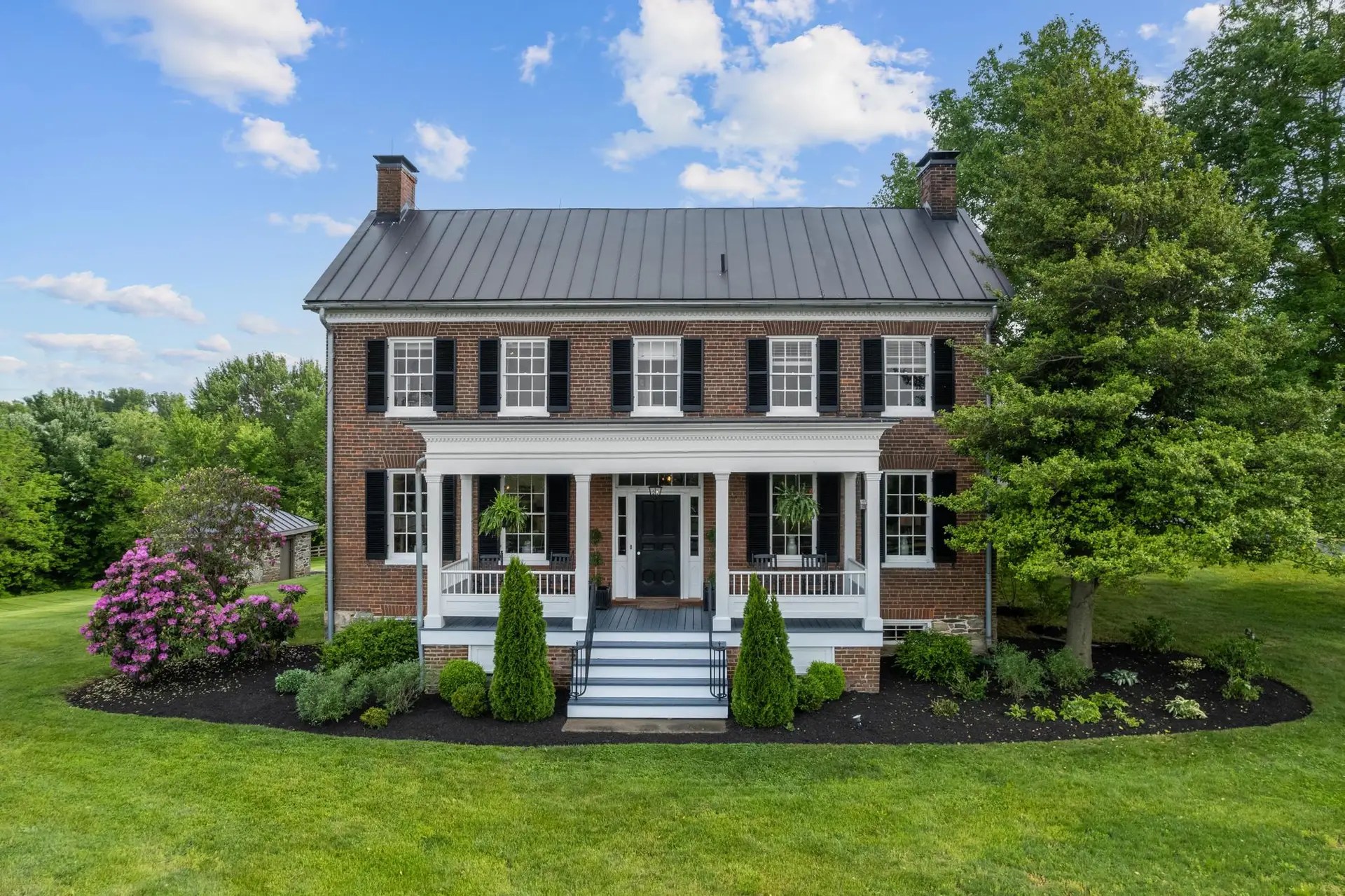 Front view of a beautiful brick colonial-style house with black shutters, a manicured lawn, and vibrant landscaping.