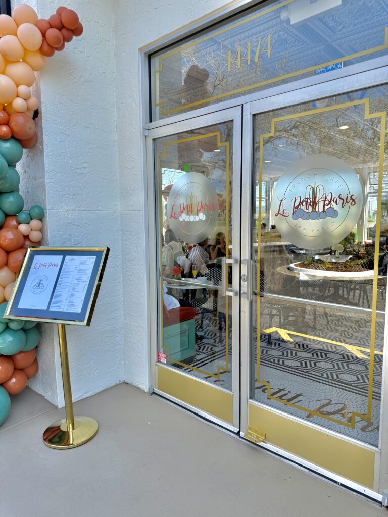 The entrance of Le Petit Paris French Café, featuring custom gold-accented vinyl logos on the glass doors and an elegant outdoor menu display.