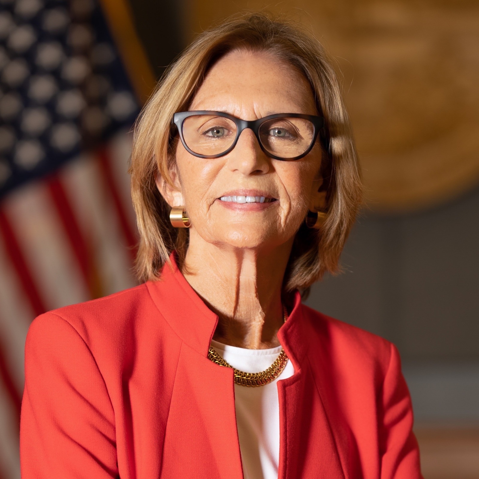 a woman in a red jacket and glasses standing in front of an american flag