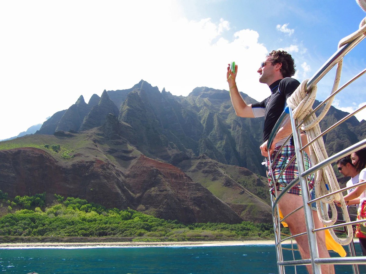 Man taking pictures of the Napali coast from the railing of a boat.