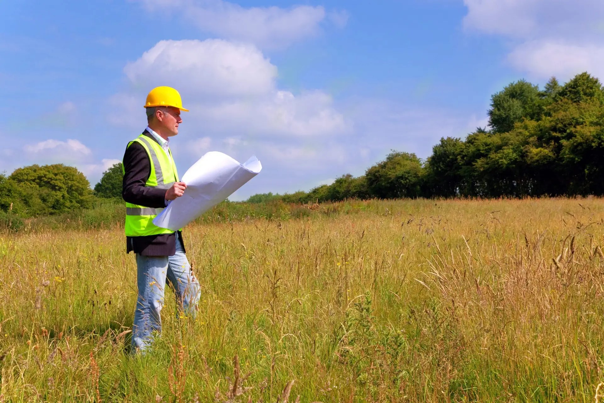 Surveyor in a high-visibility vest using equipment to assess land for a new construction project in an open field.