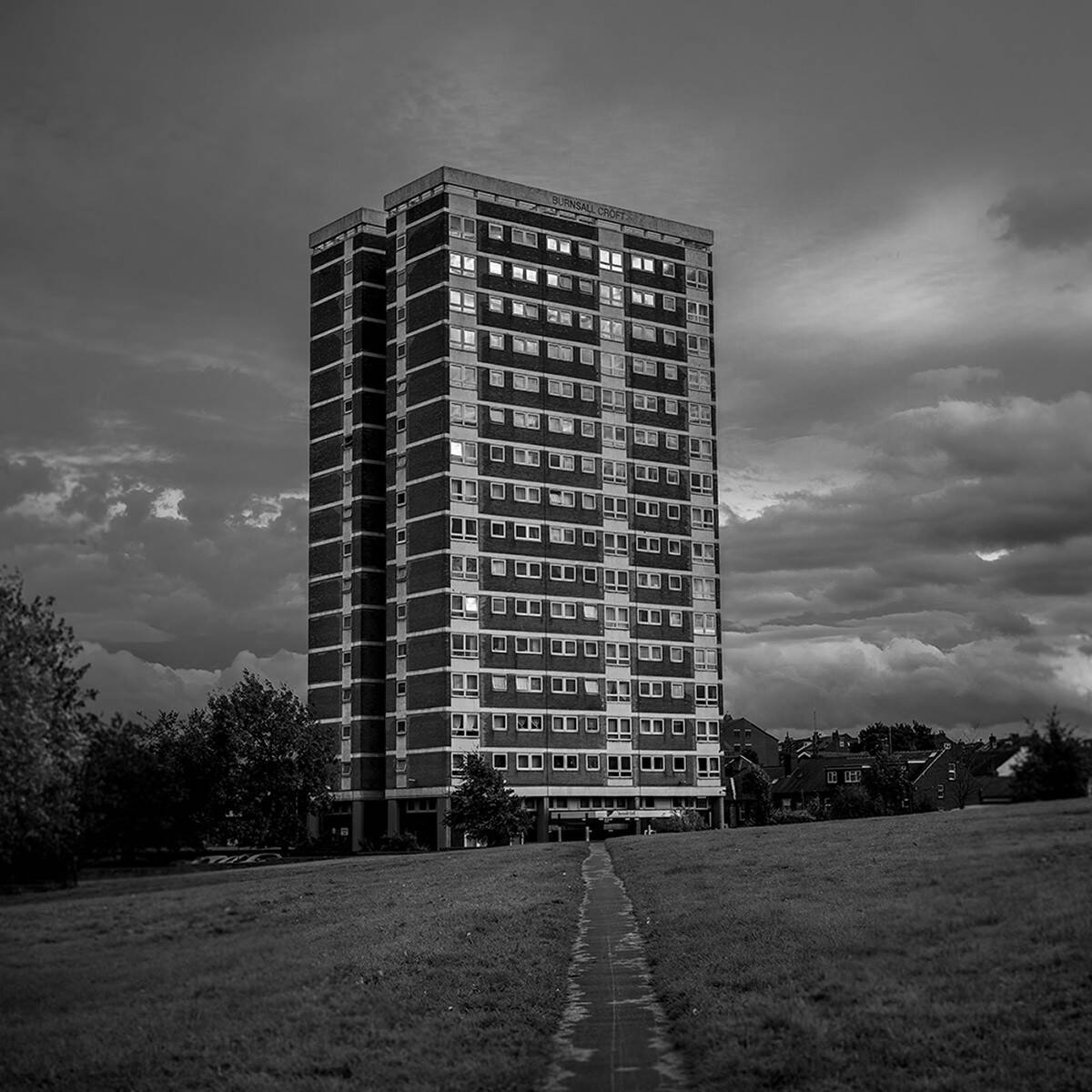 A black and white photograph portraying a moody atmosphere. A tower block stands alone on a green. A narrow path cuts through the middle leading towards the tower. The sky is cloudy and grey.