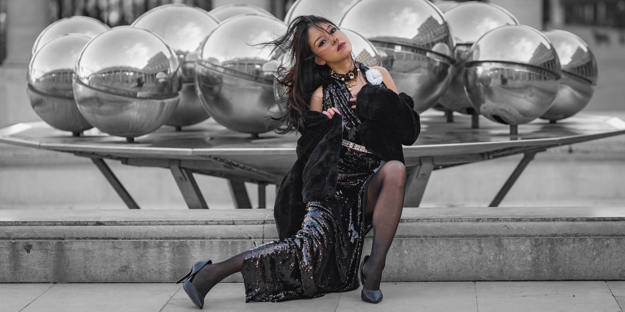 A fashion model in a black sequin dress and faux fur coat poses dynamically in front of reflective silver spheres during an on-location photoshoot collaboration.