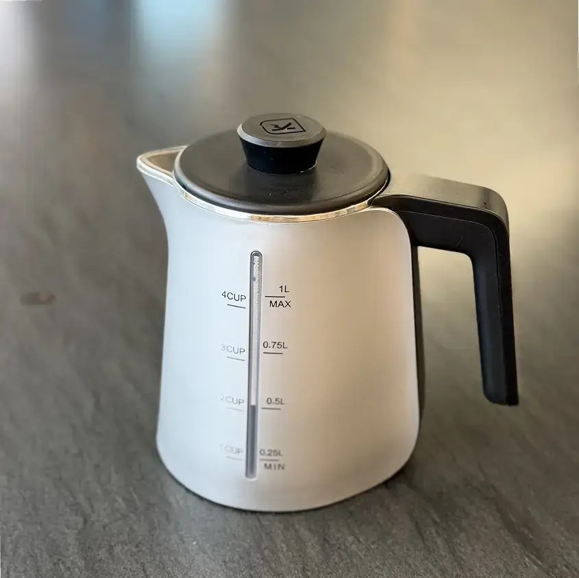 Stainless steel electric kettle with a black handle on a dark countertop, showing a vertical water level indicator and measurement marks up to 1 liter. The lid is black with a round knob.