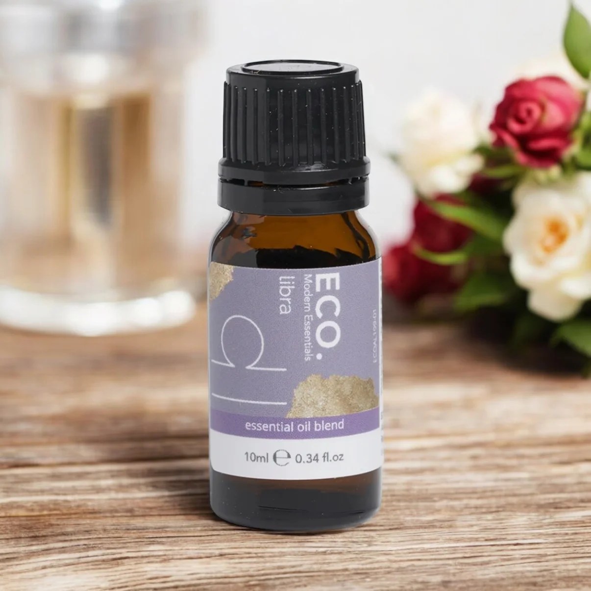 A small amber bottle of Libra Zodiac Essential Oil Blend rests on a wooden surface, with roses and a glass candle holder softly blurred in the background.