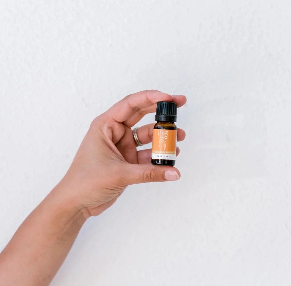 A hand with a ring holds a small amber bottle of Neroli Essential Oil 3% in Jojoba, featuring an orange label and black cap, against a plain white background.