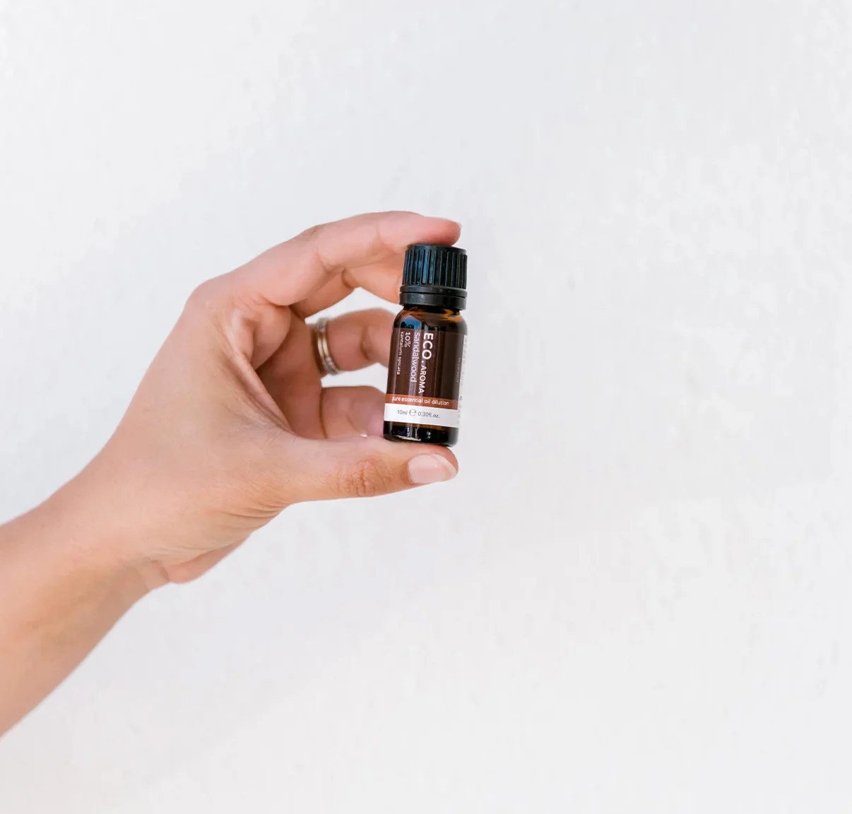 A hand with a ring on the ring finger holds a small brown bottle of Sandalwood Essential Oil 10% with a black cap and a white and red label against a plain white background.