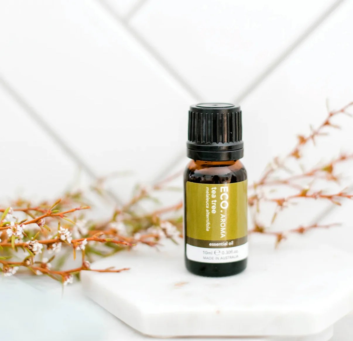 A small amber bottle of Tea Tree Essential Oil – 100% Pure Australian Melaleuca Alternifolia sits on a white marble surface, with delicate greenery in the background and a white tiled wall behind.