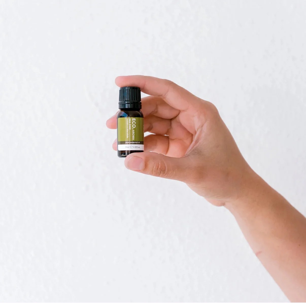A hand holds a small dark glass bottle of Tea Tree Essential Oil – 100% Pure Australian Melaleuca Alternifolia, featuring a black cap and green label, against a plain white background.