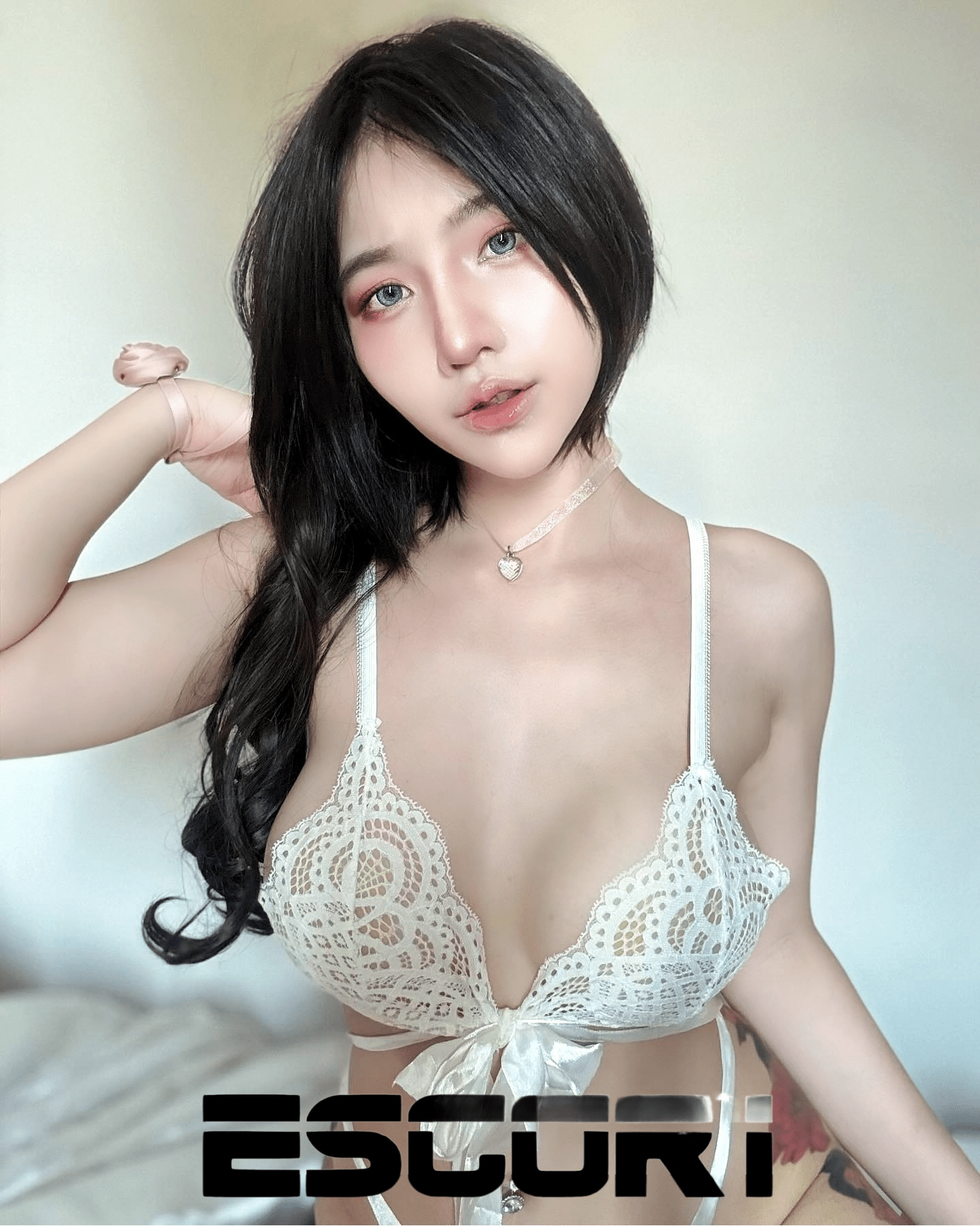 Selfie-style photo of a young woman with black hair wearing white lace lingerie, a pink scrunchie on her wrist, and a heart pendant necklace, posing softly.
