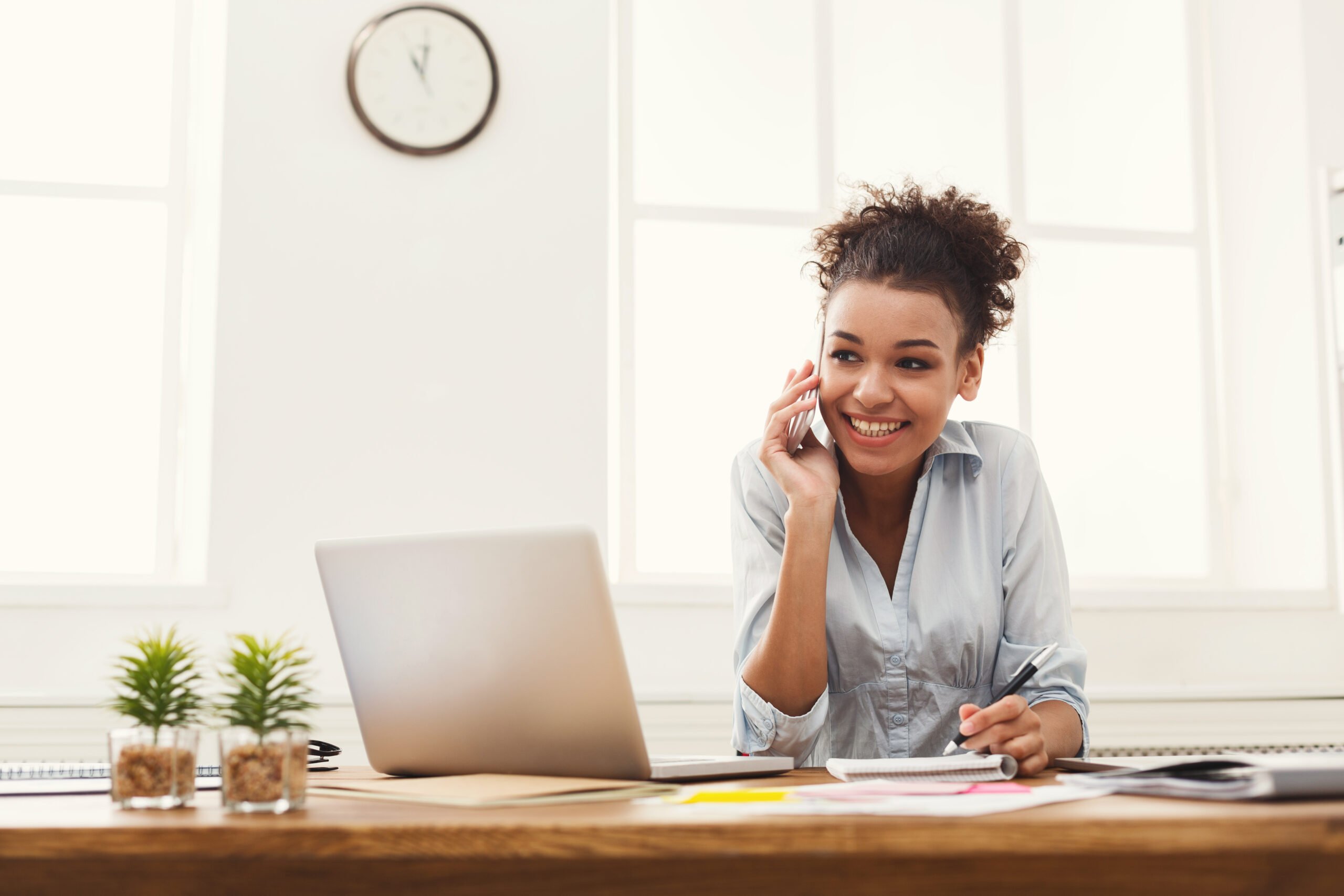 Happy smiling african-american business woman at work talking on phone and taking notes, sitting at her working place in office, copy space
