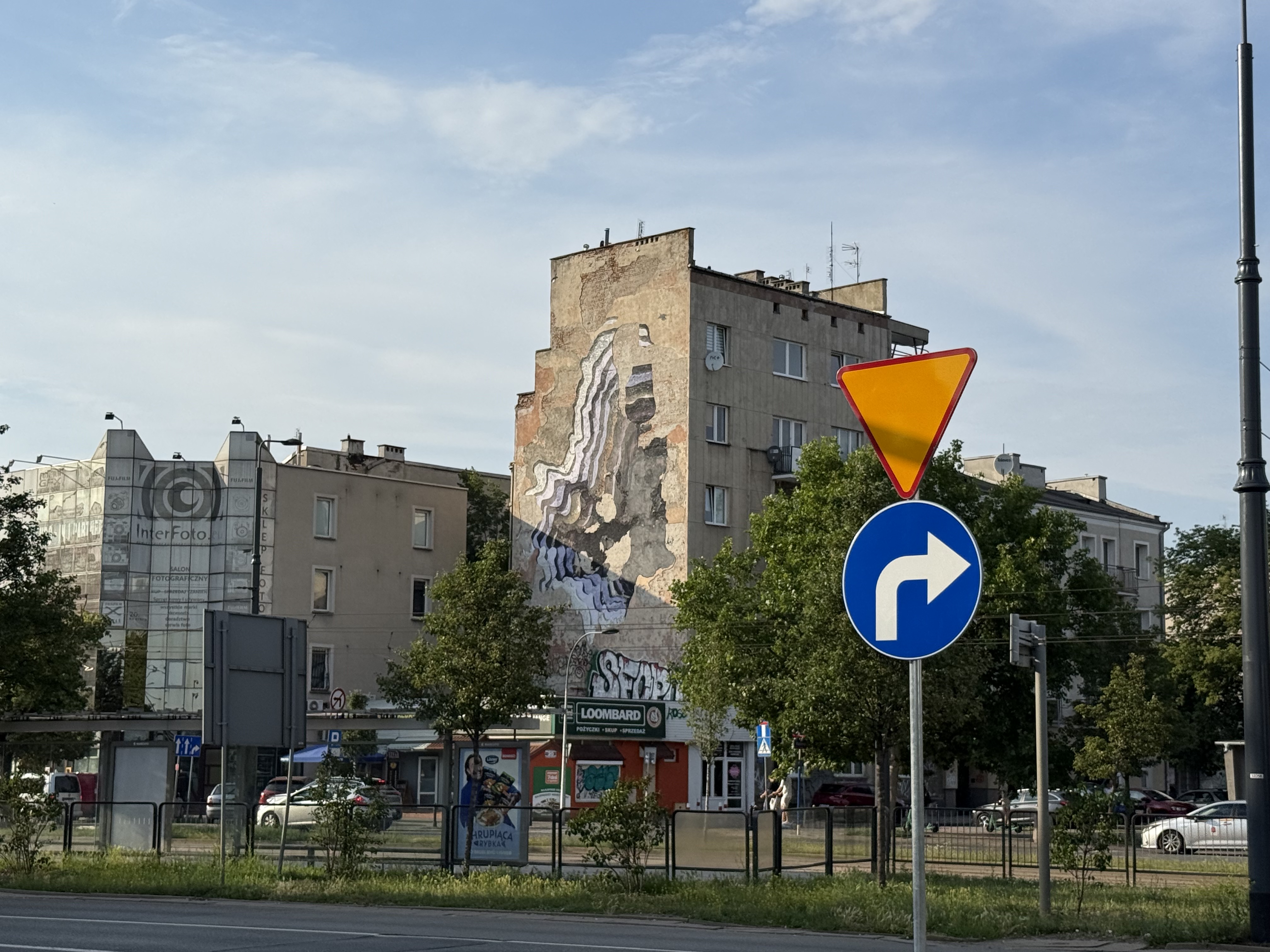 Urban street scene with a tall mural-covered building, traffic signs in the foreground, and trees along the sidewalk.