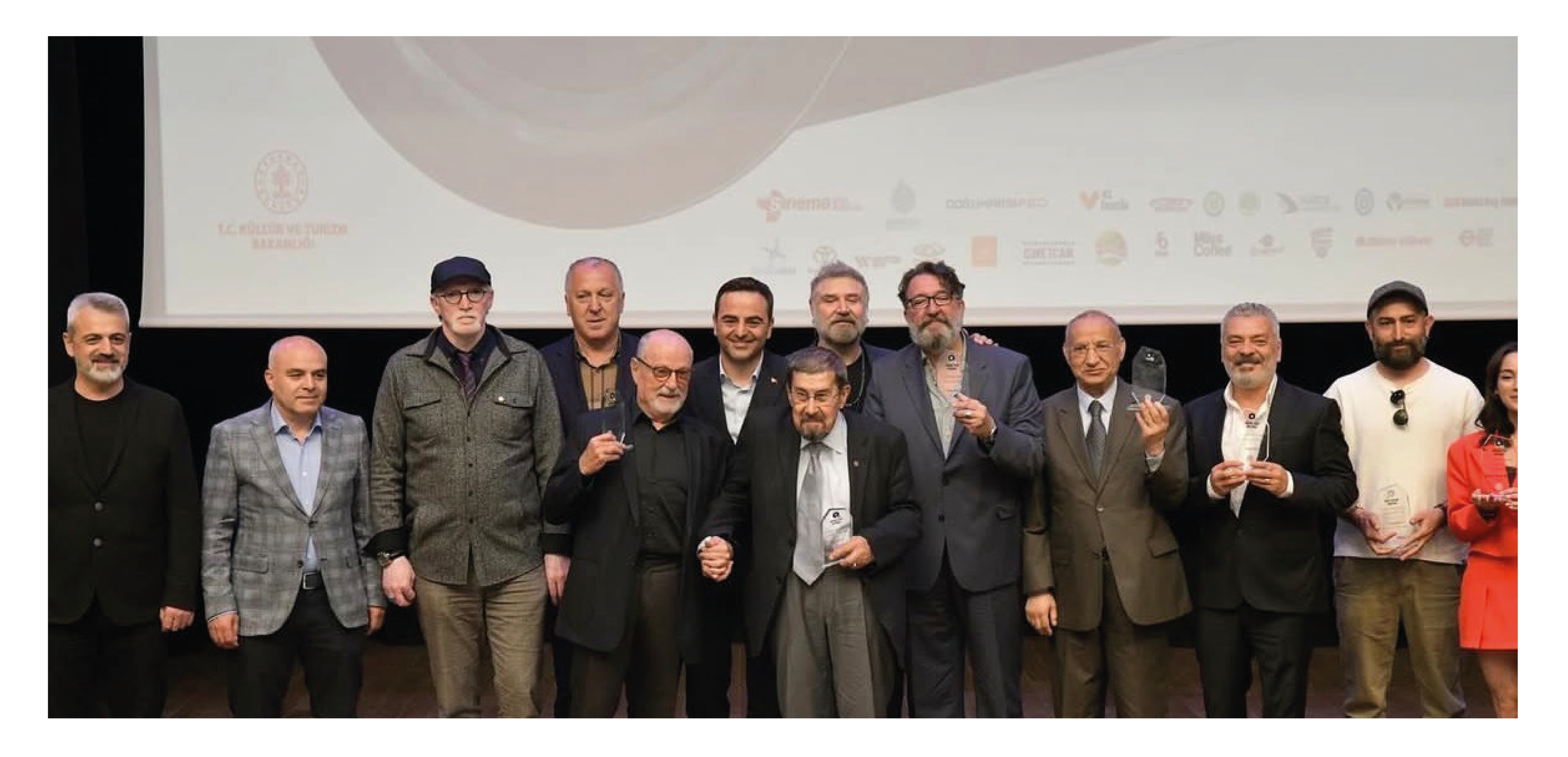 Bir ödül töreninden görüntü, sahnede çeşitli bireyler ödüllerini tutarak poz veriyor.