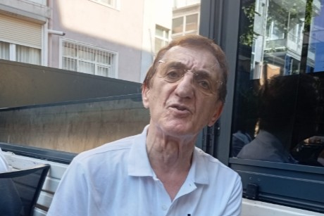 Bir kafede oturan, beyaz tişört giymiş yaşlı bir adam, kameraya bakarak konuşuyor.