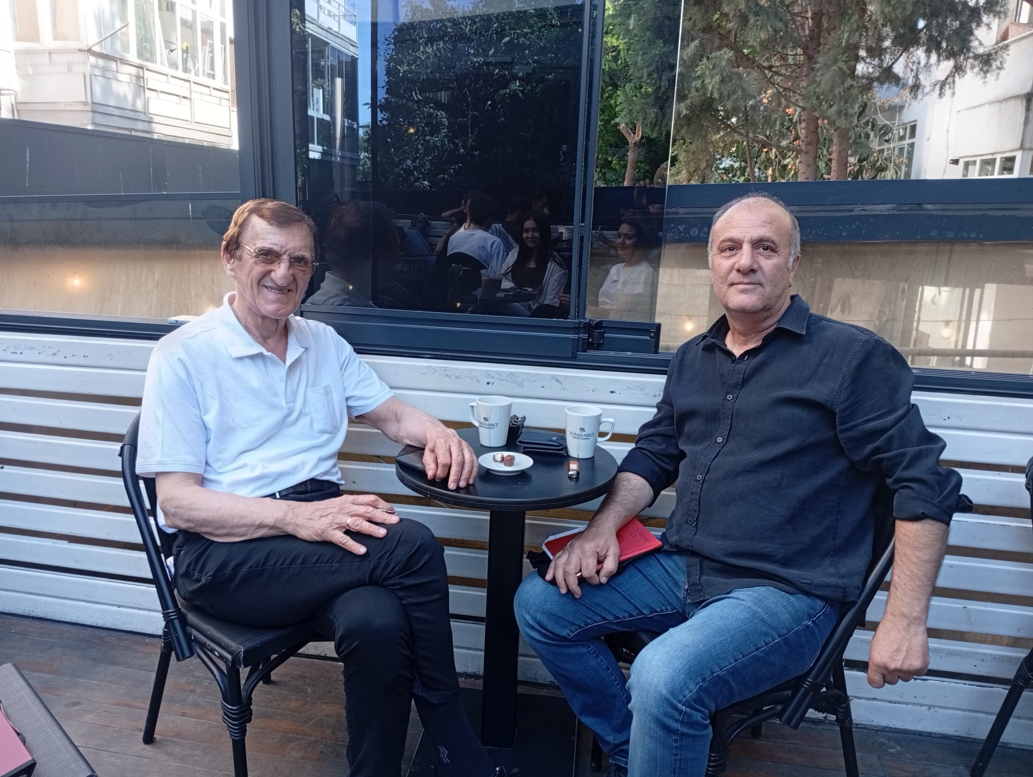 İki adam, bir kafede işlemeli masanın etrafında oturmuş, kahve içiyorlar. Arkada doğal ışıkla aydınlanmış bir ortam görünüyor.