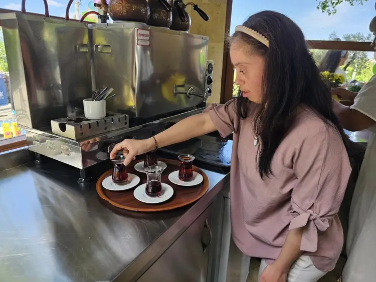 A woman serves three small glasses of tea on white plates from a wooden tray in a kitchen setting, with kitchen equipment and a bright outdoor background.