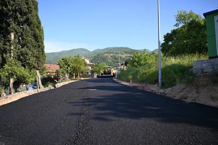Yeni asfalt serimi yapılan bir yol, yeşil ağaçlar ve dağ manzarası ile çevrili. Güneşli bir gün ve yol yüzeyine sıcak asfalt dökülüyor.