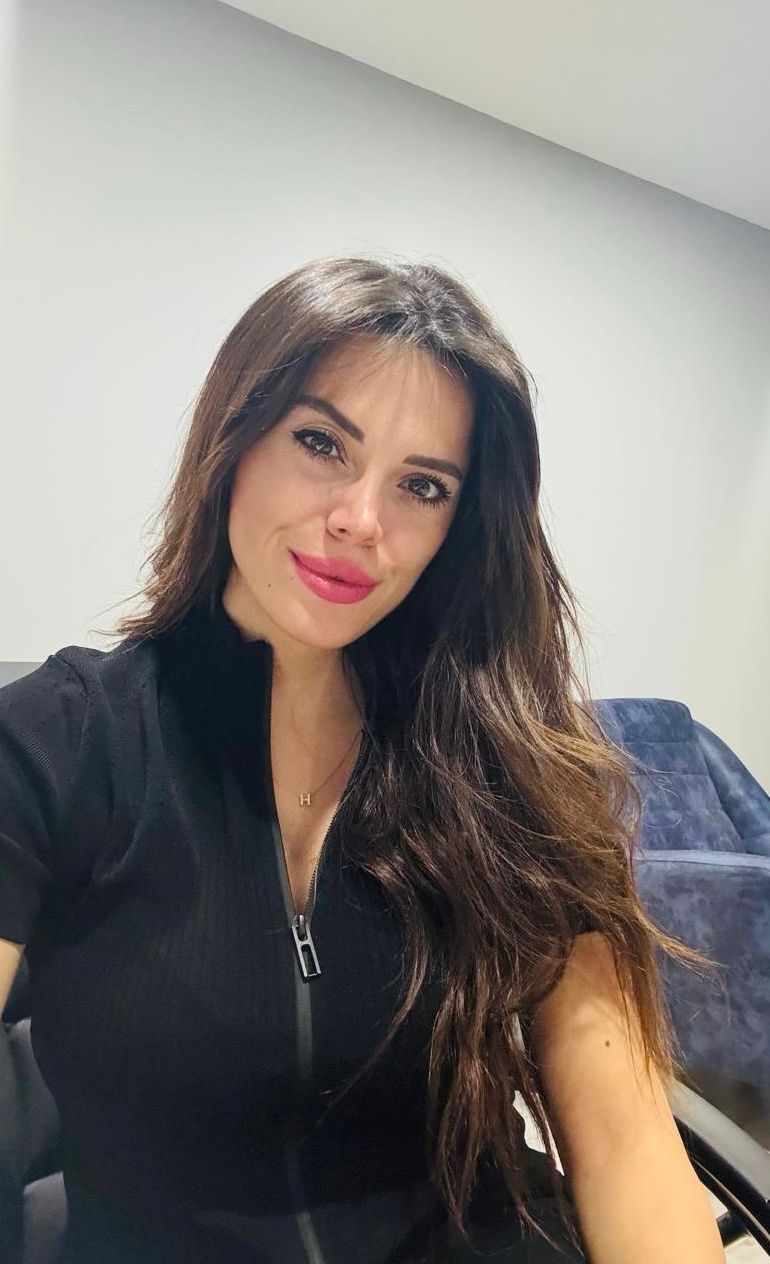 Bir kadın, siyah bluz giyerek gülümsüyor. Arka planda sade bir ofis ortamı bulunuyor.