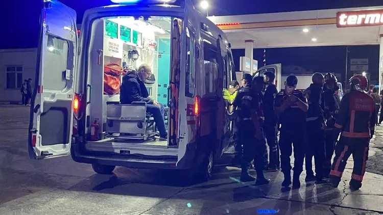 Kaza sonrası hastaneye sevk edilen yaralı bir adam ambulans içinde oturuyor. Etrafta sağlık ekipleri, polis ve itfaiye personeli bulunuyor. Arka planda bir akaryakıt istasyonu görünmekte.