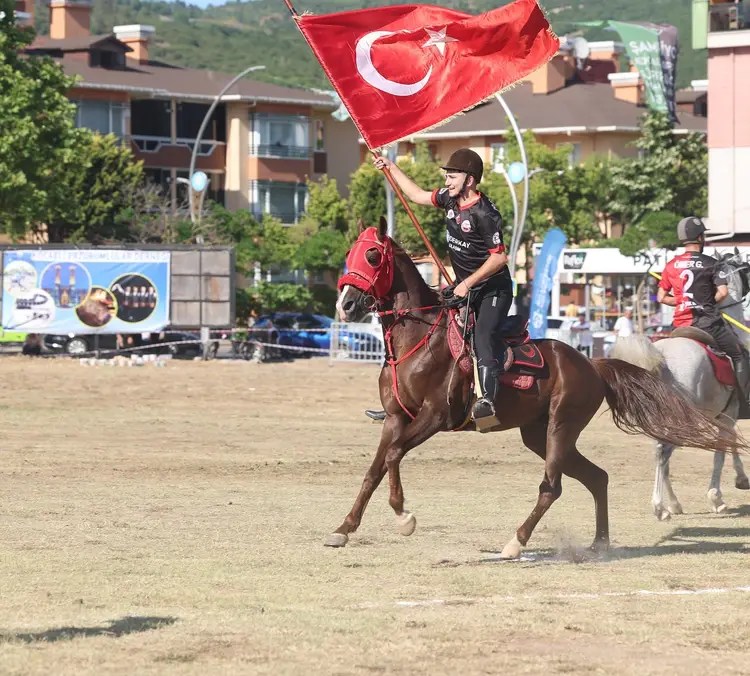 Bir at üzerinde, Türk bayrağını dalgalandıran bir kişi. Etkinlik alanında diğer atlı sporcular yer alıyor.