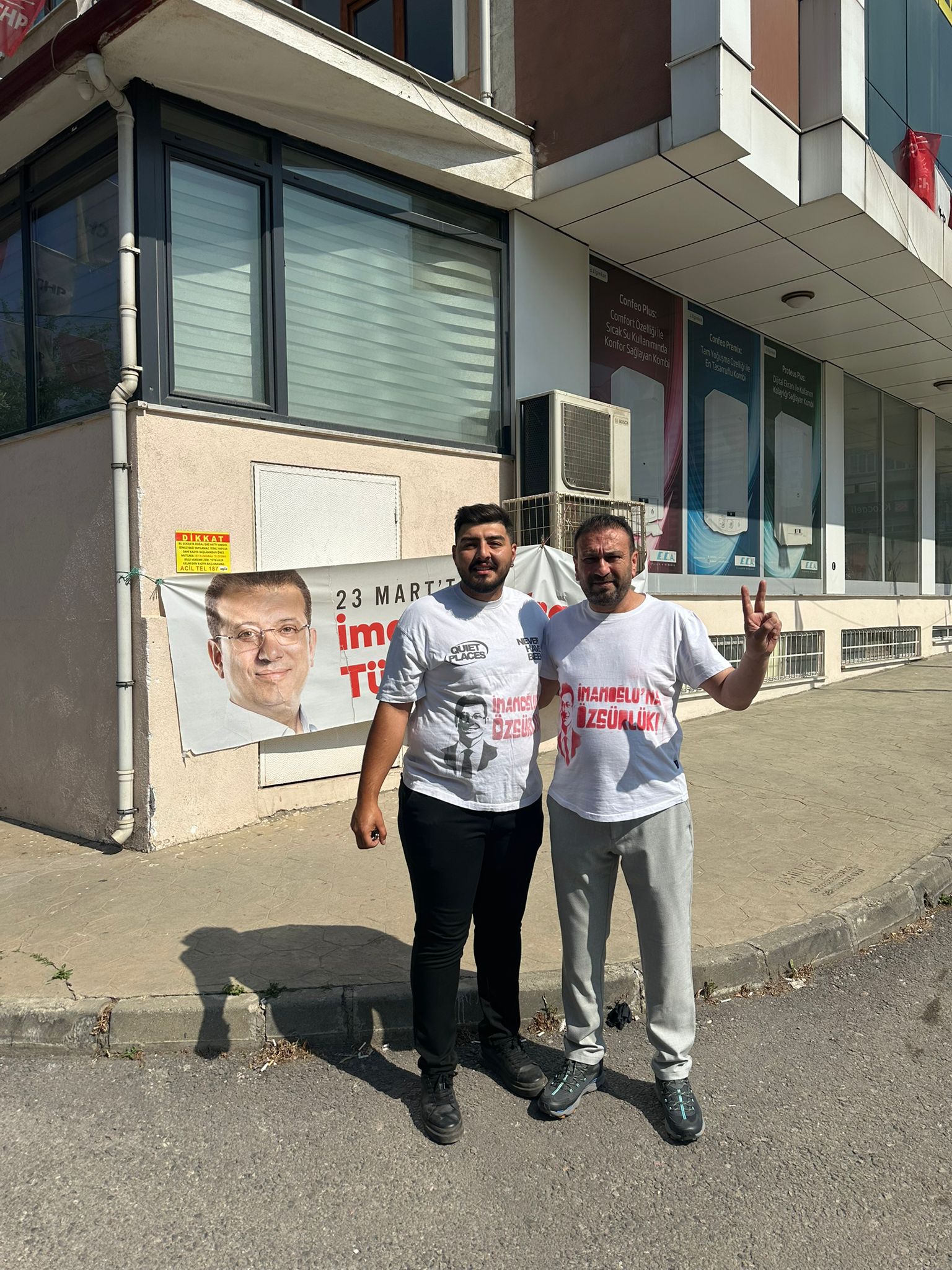 İki genç adam, 'Adalet, hak, hukuk için İmamoğlu'na özgürlük' yazılı tişörtlerle poz veriyor. Arkada bir binanın duvarında İmamoğlu'nun resmi ve protesto afişleri görülüyor.