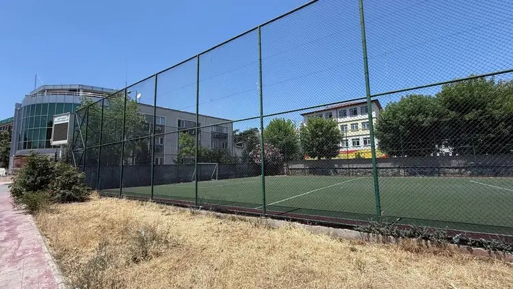 Halı saha görüntüsü, etrafında yeşil alanlar ve binalar mevcut. Ağaçlar ve çalılarla çevrili, sahada boş bir alan görünüyor.