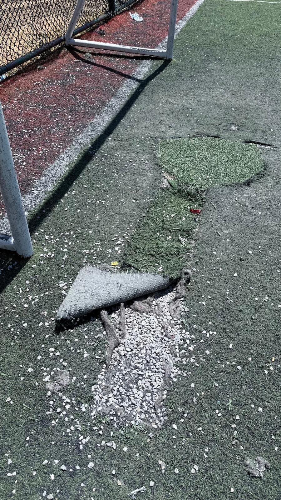 Yıpranmış ve bakımsız bir futbol sahasında zayıf onarımların yapıldığı bir alan. Zemin üzerinde parçalanmış halı ve taşlar görünmekte.