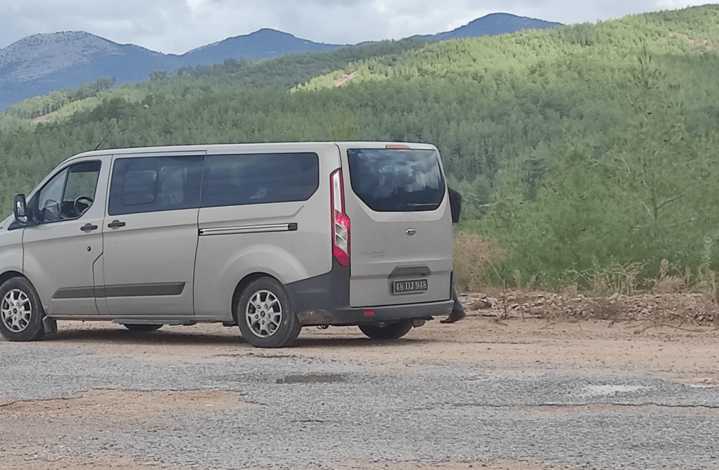 Gri bir minibüs, dağlık ve yeşil bir alanda duruyor.