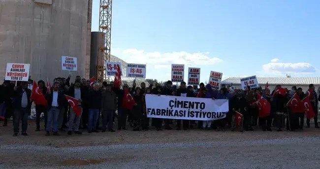 Protesto gösterisi sırasında bir grup insan, 'Çimento Fabrikası İstiyoruz' yazılı pankart tutuyor ve Türk bayrakları taşıyor.